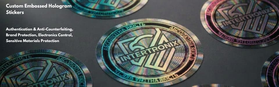Multiple iridescent embossed holographic stickers bearing the Intertronix logo are arranged on a dark surface, their rainbow diffraction patterns catching studio light, showcasing custom hologram authentication and brand protection solutions.